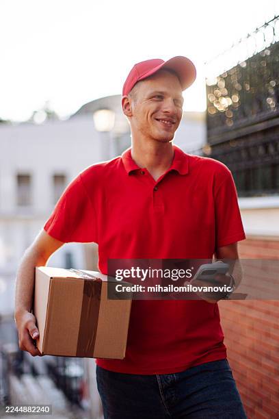 repartidor con polo rojo y gorra sonriendo mientras sostiene la caja y el teléfono inteligente al aire libre - entrega sin contacto fotografías e imágenes de stock