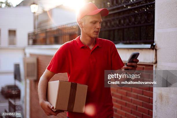 repartidor en polo rojo revisando el teléfono mientras lleva la caja al aire libre en un entorno urbano - entrega sin contacto fotografías e imágenes de stock