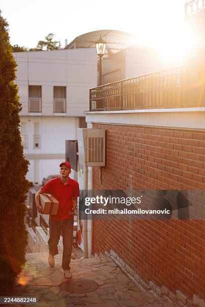 repartidor llevando paquete a lo largo de la pared de ladrillos iluminada por el sol en callejón urbano al atardecer, hora dorada - entrega sin contacto fotografías e imágenes de stock