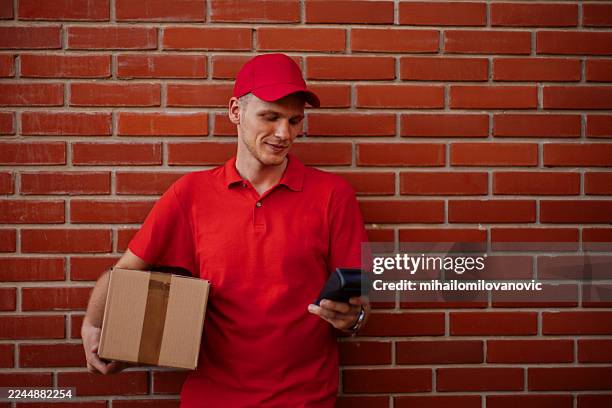 repartidor con uniforme rojo de pie junto a la pared de ladrillos usando el teléfono mientras sostiene el paquete - entrega sin contacto fotografías e imágenes de stock