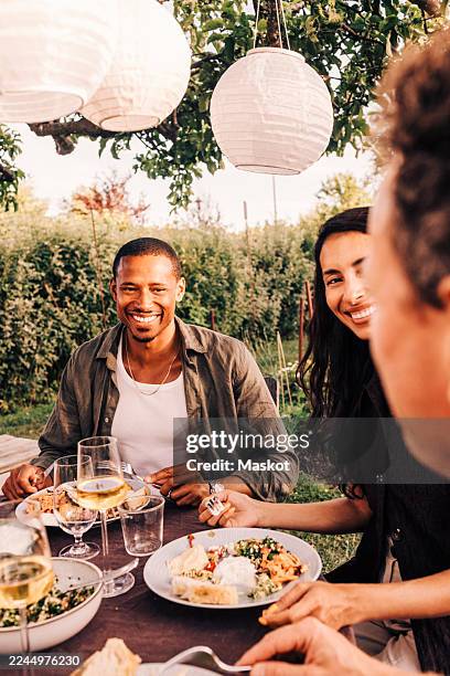 smiling man eating food while enjoying with family friends at dinner party in back yard - lampion photos et images de collection