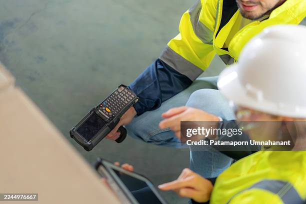 two warehouse workers wearing protective workwear are scanning barcodes with a barcode reader and verifying product data on a digital tablet in a large distribution warehouse. the scene shows teamwork, accuracy, and modern inventory management - radio frequency identification stock pictures, royalty-free photos & images