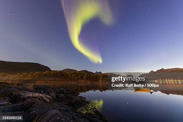 stunning aurora borealis dances over a tranquil lake with a lone boat at night, reflecting the vibrant green and purple lights in the water,senja,norway,fjordbotn camping - insel senja stock-fotos und bilder