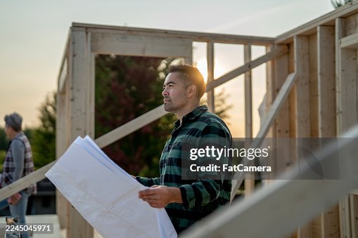 Asian Man Construction Worker Analyzing Blueprints On Outdoor Building Site At Sunset With Plans And Drafts Asian Man Construction Worker Analyzing Blueprints On Outdoor Building Site At Sunset With Plans And Drafts