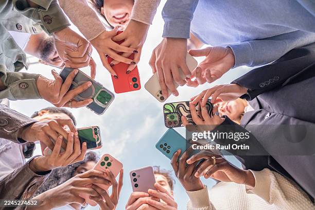 group of young people using smartphones, forming a circle and looking down at their devices - acoso escolar fotografías e imágenes de stock