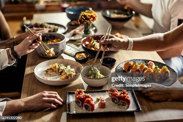a table full of various asian dishes, with sushi and other small bowls of side dishes dominating the foreground. several hands with chopsticks are in the shot, suggesting active conversation and shared enjoyment of the meal in the restaurant. - japanese food stock pictures, royalty-free photos & images