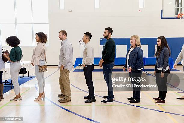 diverse group of adults stand patiently in line inside a gymnasium, awaiting their turn. - stembureau stockfoto's en -beelden