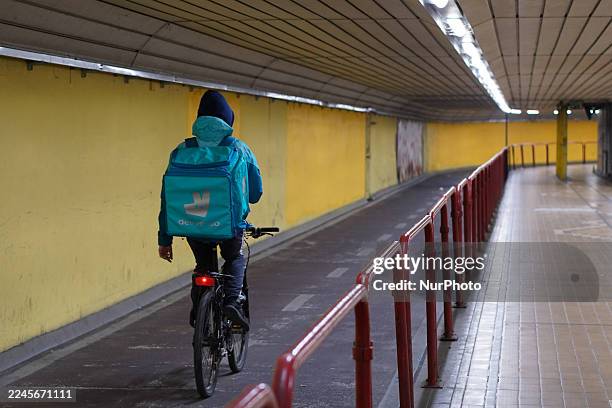 Food courier wears a bright blue jacket and a large Deliveroo backpack and rides a bicycle through a brightly lit underpass or pedestrian tunnel in...