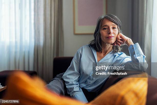 thoughtful woman relaxing on sofa in casual blue shirt in modern living room setting - mulheres maduras imagens e fotografias de stock