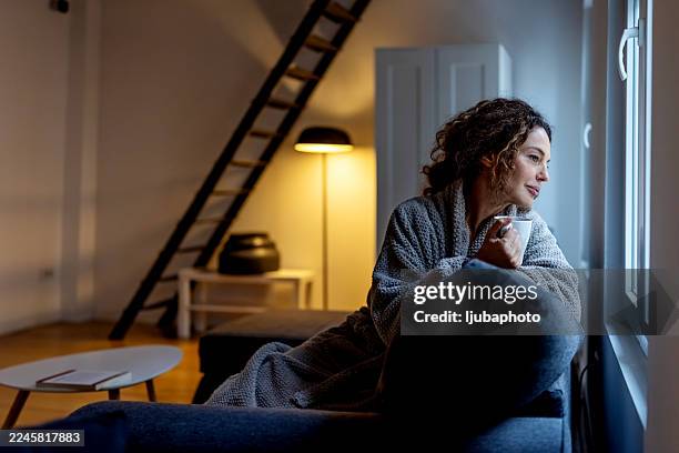ambiente acogedor por la noche: mujer relajándose junto a la ventana con una taza en el salón suavemente iluminado - penumbra fotografías e imágenes de stock