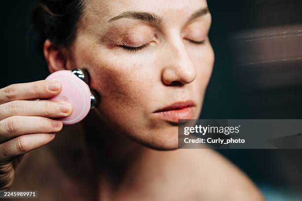 a close-up of a middle-aged woman with closed eyes applying a small, pink facial massage device to her cheek, highlighting her natural skin texture and freckles against a dark background. - massaggio del viso foto e immagini stock