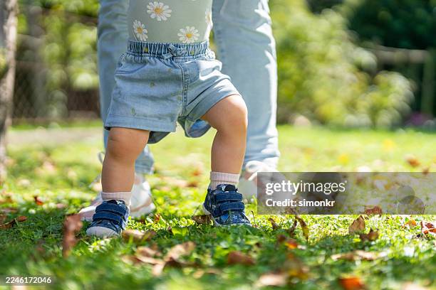 joyful young caucasian girl learning to walk with mother's guidance in sunlit garden - baby clothing stock pictures, royalty-free photos & images