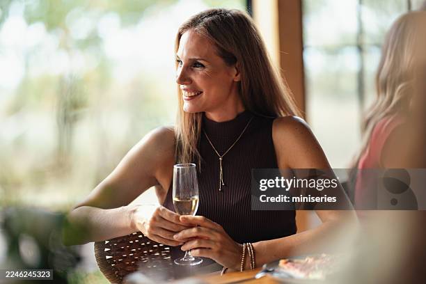 lächelnde frau, die champagner am esstisch in einem hellen restaurant während eines entspannten mittagessens genießt - essen im freien stock-fotos und bilder