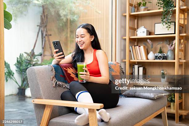 young asian sports woman resting and having green smoothie after exercising at home in the morning, using smartphone - achatar la curva de epidemia fotografías e imágenes de stock