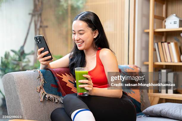 young asian sports woman resting and having green smoothie after exercising at home in the morning, using smartphone - aplatir la courbe photos et images de collection