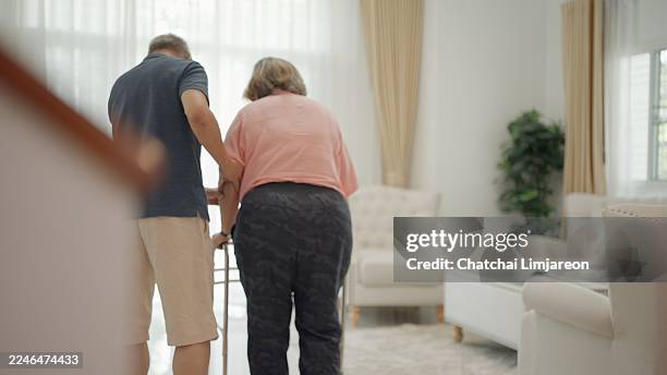 rear view of husband takes care of his wife, who is exercising with crutches, and then sits together in the living room to recuperate from an illness at home. retired couple takes care of each other's health. retirement activities, lifestyles, well-being. - crutch stock pictures, royalty-free photos & images