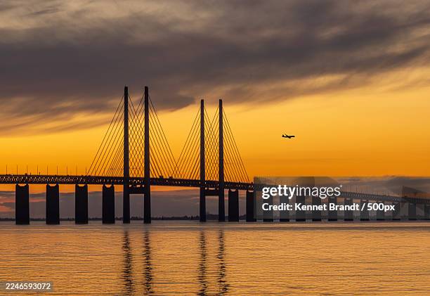 silhouette of a majestic bridge over calm waters against a vibrant sunset sky with an airplane flying overhead,sweden - malmo stock pictures, royalty-free photos & images