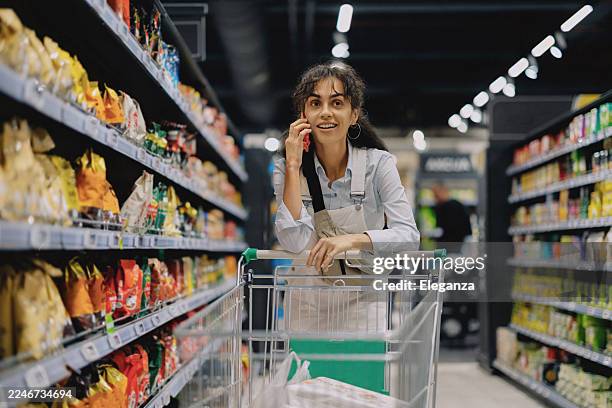 jeune femme poussant un caddie dans un supermarché tout en vérifiant sa liste d’épicerie sur son smartphone. elle fait ses courses hebdomadaires, choisit des produits alimentaires, des produits d’entretien ménager et des articles de soins personnel - produit dentretien photos et images de collection