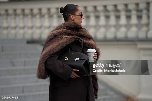 Anna Wein is seen wearing a full lenght Bazhane coat in brown with a straight, longline cut with a slightly oversized drape; an oversized brown fur...