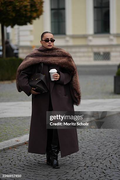 Anna Wein is seen wearing a full lenght Bazhane coat in brown with a straight, longline cut with a slightly oversized drape; an oversized brown fur...