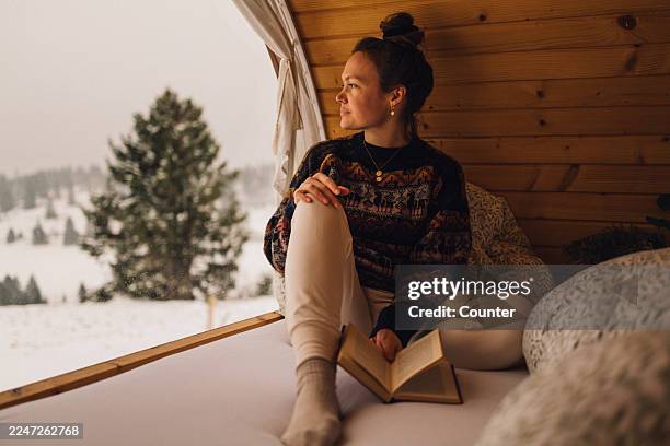 a woman reading a book inside a warm, cozy wooden winter cabin - cozy winter stock pictures, royalty-free photos & images