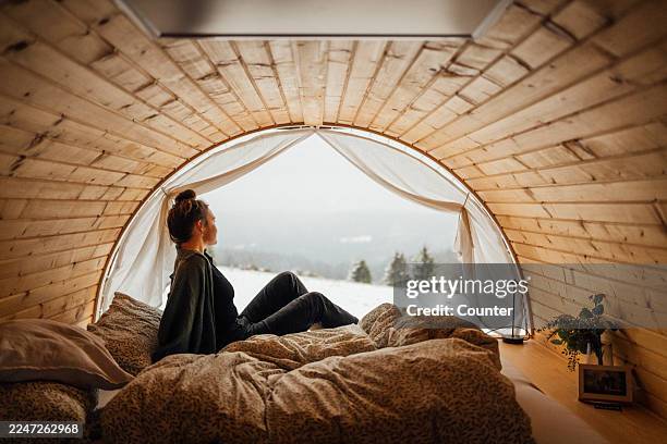 cozy winter cabin bedroom with woman looking out over snowy landscape - glamping stock-fotos und bilder