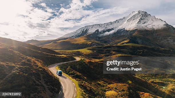 an aerial/drone view of a blue campervan driving on a mountain pass in the french pyrenees. the road winds towards a snowcapped mountain - dramatic landscape stock pictures, royalty-free photos & images