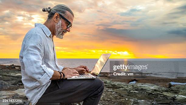 man with glasses working on laptop by the sea at sunset - digital nomad stock pictures, royalty-free photos & images