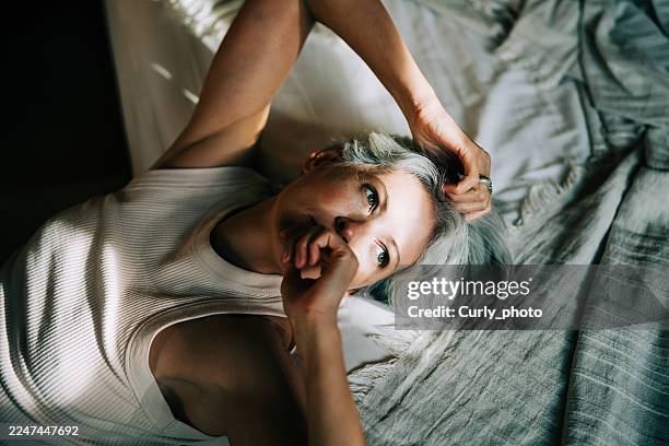 a middle-aged woman with gray hair lies on her back in bed, illuminated by bright sunlight and shadow. - hormone photos et images de collection