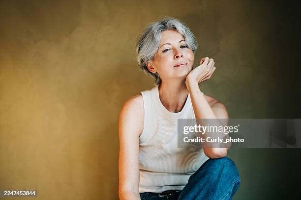 a cheerful middle-aged woman with short gray hair against a warm, textured gold wall, smiling genuinely at the camera. - grijs haar stockfoto's en -beelden