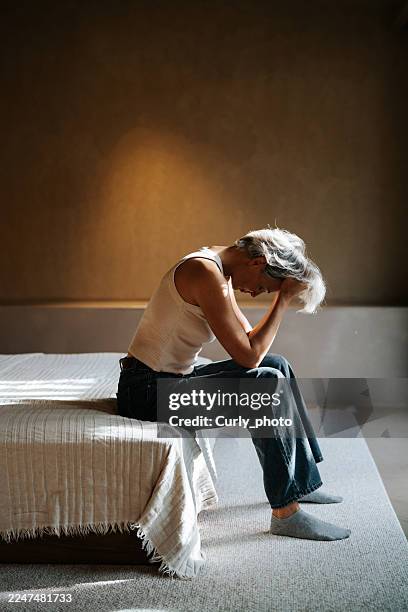a middle-aged woman with gray hair sits on the edge of a bed in a dark room, her head in her hands. this powerful, oppressive scene conveys the stress, anxiety, and emotional exhaustion of a moment of personal crisis or isolation. - ormone foto e immagini stock