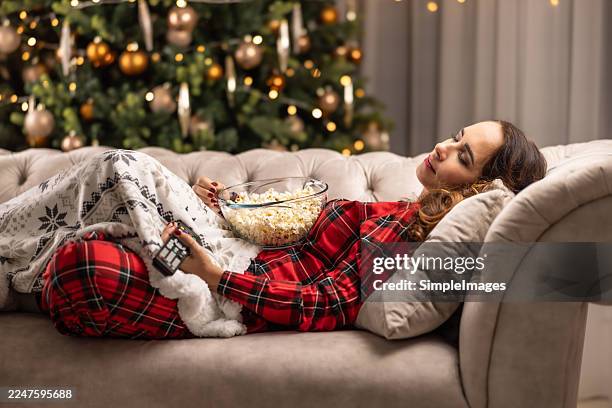 the christmas cheer has finally overcome the fatigue, and a woman in pajamas is sleeping on the couch under a blanket, with a bowl of popcorn and a remote control next to her, while a christmas tree glows in the background. - christmas television stock pictures, royalty-free photos & images