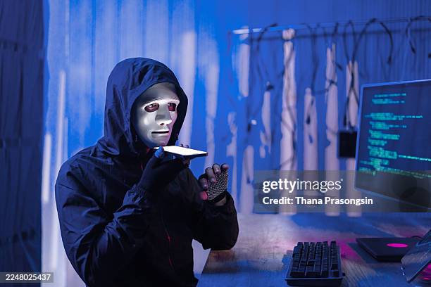 recording a voice message on a smartphone while sitting at a desk with a computer displaying code. - scammer stock pictures, royalty-free photos & images