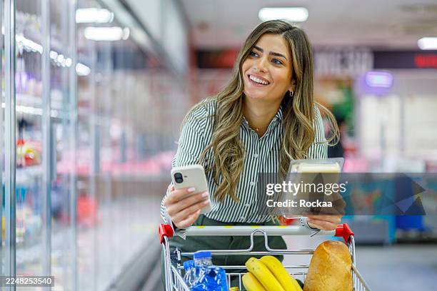 happy woman using smartphone and shopping cart while looking for products in supermarket freezer aisle - mand stockfoto's en -beelden