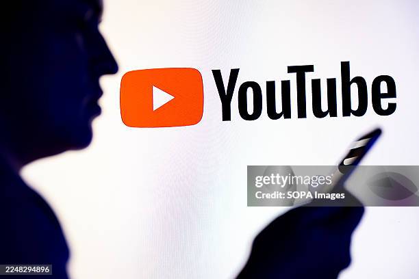 In this photo illustration, a silhouetted individual is seen holding a mobile phone with a YouTube logo displayed in the background.