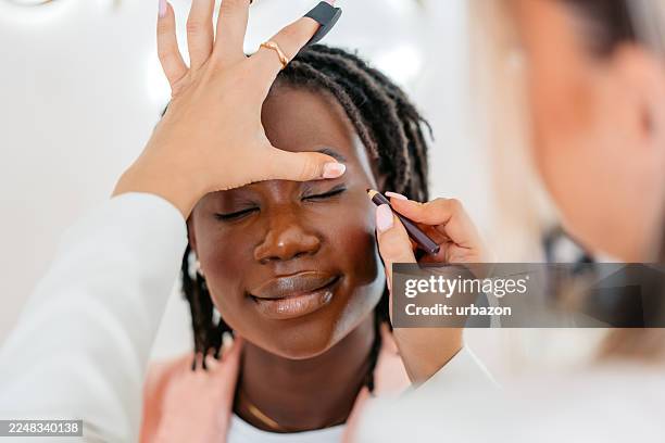 kosmetikerin, die eyeliner auf eine junge schwarze kundin in einem schönheitssalon aufträgt - permanent make up stock-fotos und bilder