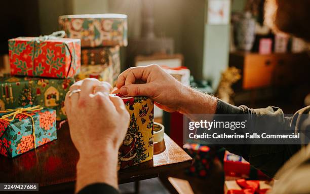 a scene of gift wrapping at christmas, using bright, vintage style christmas wrapping paper. a man tucks the edges of the wrapping paper neatly as he finishes covering the present. - pinching stock pictures, royalty-free photos & images