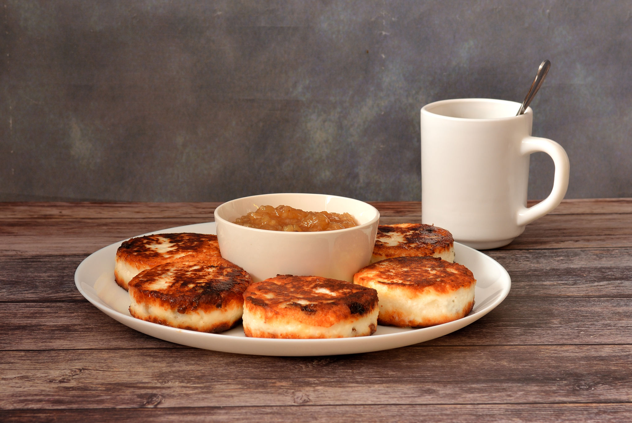 A large plate of freshly fried cheesecakes with raisins and apple jam on a wooden table, a cup of tea nearby. Close-up.