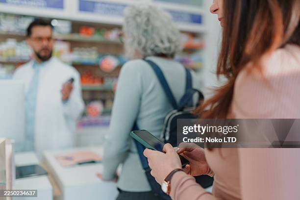 customer checking smartphone while waiting in pharmacy queue - store queue stock pictures, royalty-free photos & images