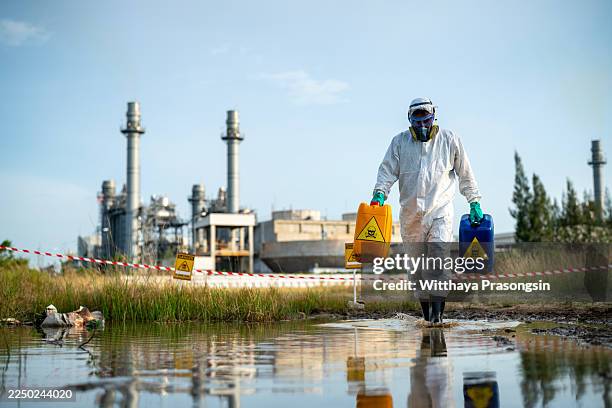 person in hazmat suit carrying hazardous waste containers near industrial facility - protective suit stock pictures, royalty-free photos & images