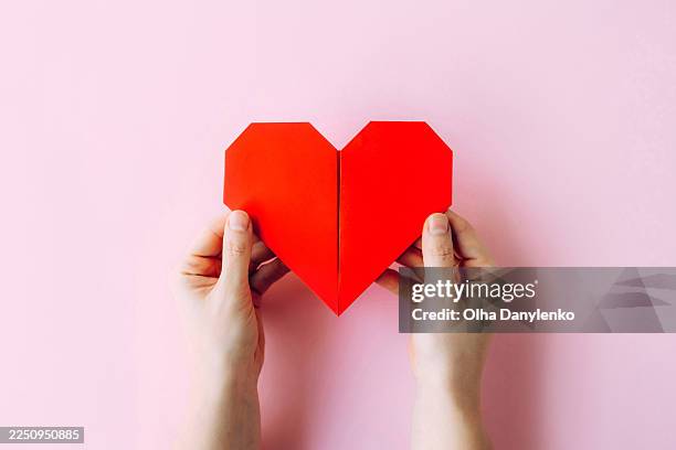 close-up of two hands holding a red paper heart on a pastel pink background with copy space. - altruismus stock-fotos und bilder
