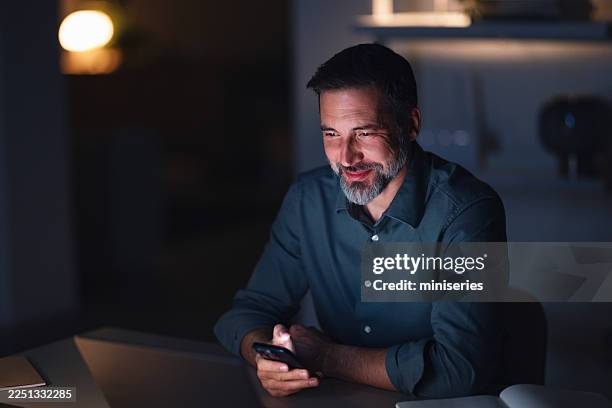 businessman working late at night in modern office, smiling while using phone - chiaroscuro stock pictures, royalty-free photos & images