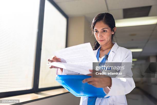 female hispanic doctor looking over medical test results in a hospital hallway - cardiologist stock pictures, royalty-free photos & images