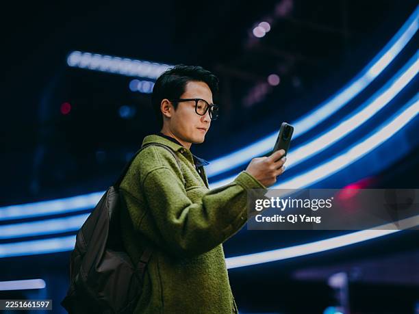 confident asian man using a smartphone while standing under an illuminated pedestrian bridge. - wealth management technology stock pictures, royalty-free photos & images