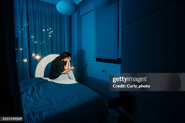 a woman sitting on the edge of a bed in a dimly lit blue room, illuminated by a soft spotlight and the glow of a smartphone. - temas de mujeres fotografías e imágenes de stock