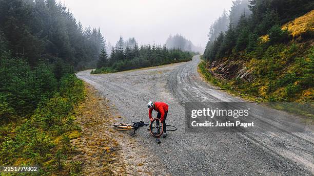 an elevated/drone view of a mature man changing an inner tube on the wheel of his gravel bike whilst on a gravel trail through forest in scotland - piercing stock pictures, royalty-free photos & images
