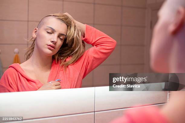 young woman suffering from cancer putting wig on her head in front of the bathroom mirror - wig stock pictures, royalty-free photos & images