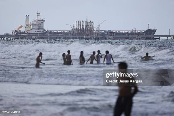 People bathe in the sea as an oil tanker is seen anchored at the dock in the outskirts of 'El Palito' refinery on December 18, 2025 in Puerto...
