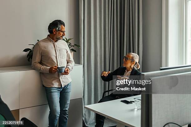 senior businesswoman discussing with male colleague while sitting on chair in office - lässig schicker stil stock-fotos und bilder