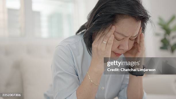 woman sits alone in her quiet, familiar home, her hands clutching her temples. her face shows the pain of a severe headache, possibly caused by stress or anxiety, panic attacks, abnormal blood pressure, or neurological condition or chronic health problem - dizzy stock pictures, royalty-free photos & images